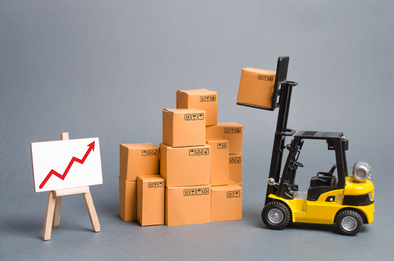 A toy forklift lifting a cardboard box, with a stack of boxes and a board showing a rising red graph – symbolizing logistics growth or price increase.