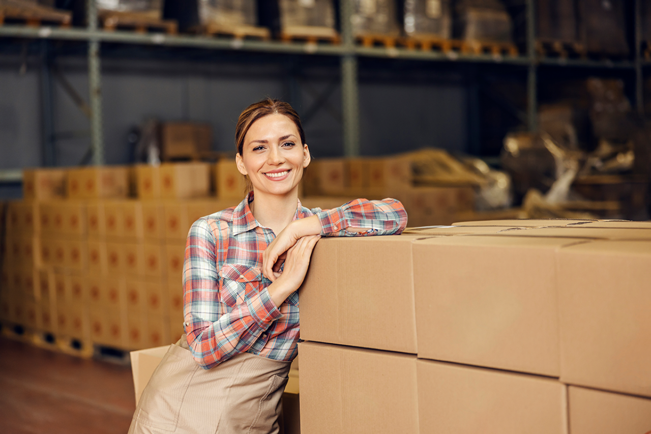 A woman smiling and leaning on shipping boxes in a warehouse.