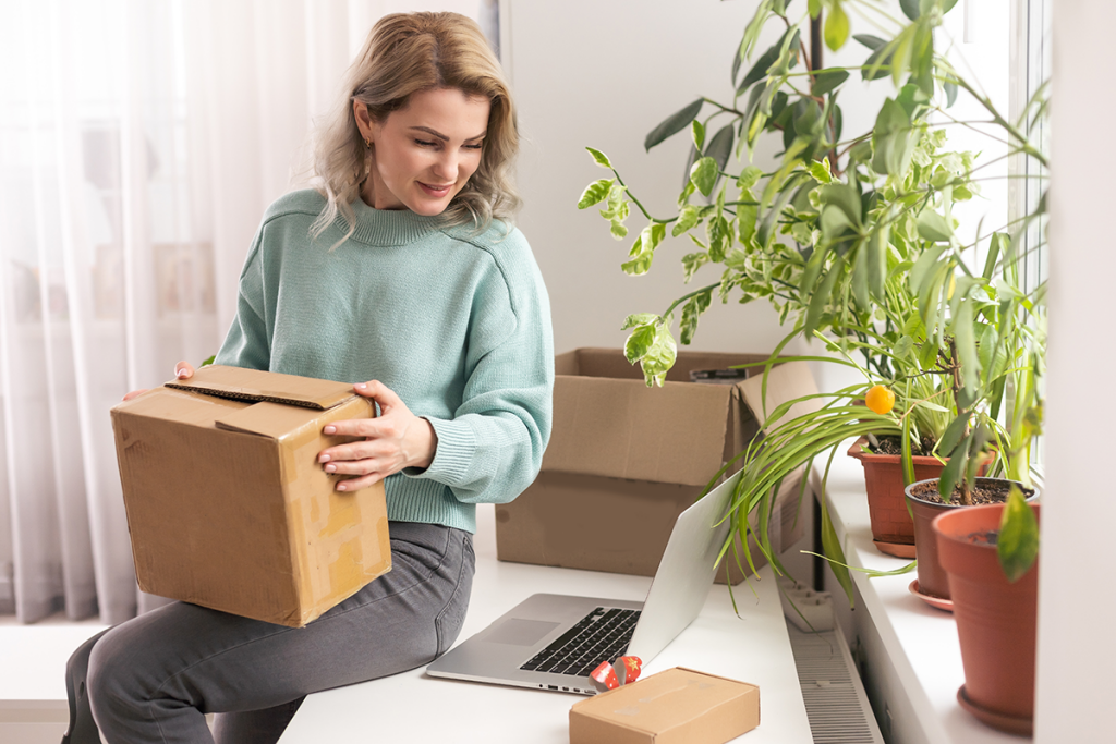 A woman is sitting on a table holding a brown package. Next to her is a laptop, and several green plants are on the windowsill.