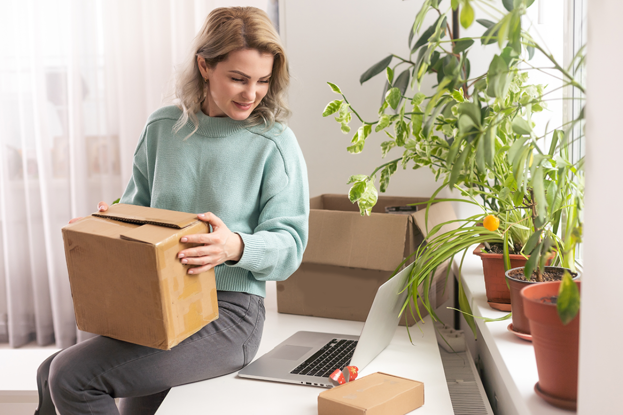 A woman is sitting on a table holding a brown package. Next to her is a laptop, and several green plants are on the windowsill.