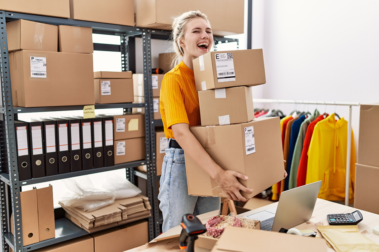 A smiling woman is carrying several packages in a warehouse with shelves, clothing, and a desk with a laptop.