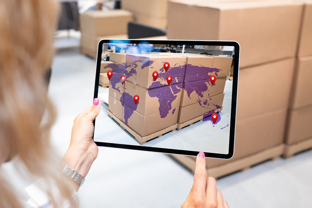 A woman is viewing brown packages through a tablet screen, with a purple world map and red location markers projected onto the boxes.