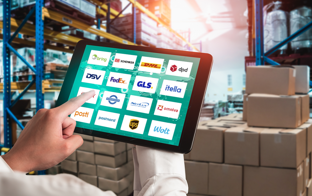 Henkilö käyttää tablettia varastossa, ruudulla näkyy useiden kuljetus- ja logistiikkayhtiöiden logoja, kuten Posti, DHL, GLS, UPS ja Wolt.