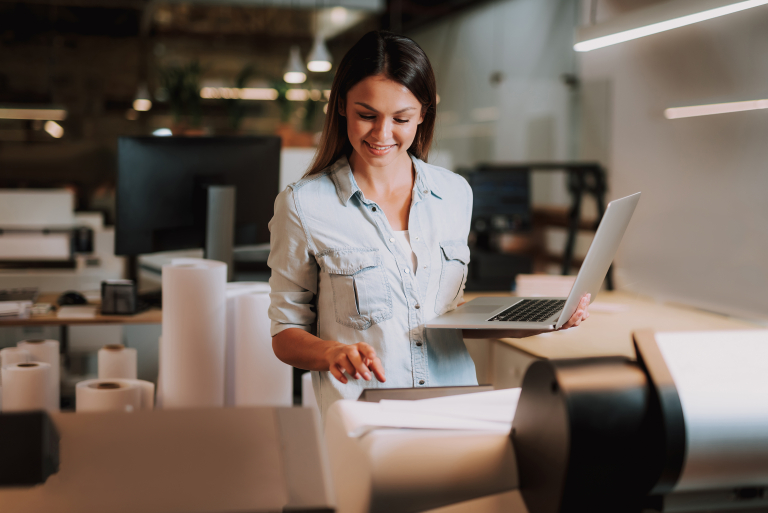 Nainen seisoo toimistoympäristössä, käyttää suurta tulostinta ja pitää kannettavaa tietokonetta toisessa kädessään.