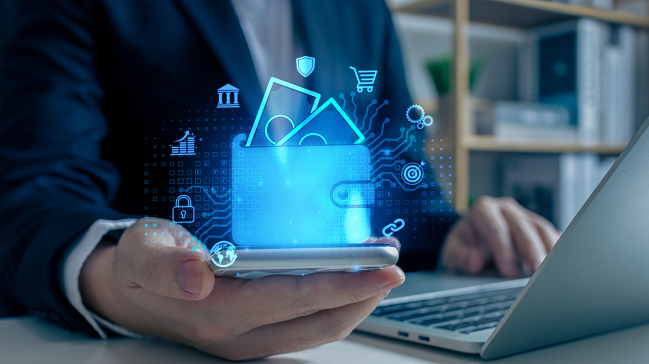 Liikemies pitää kädessään älypuhelinta, josta nousee sininen digitaalinen lompakko ja maksamisen symboleja – kuva esittää sähköisiä maksutapoja.