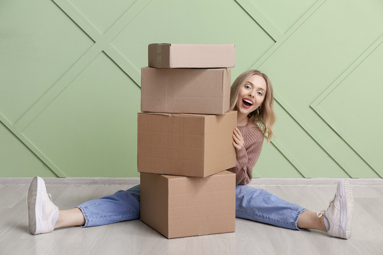 Iloinen nainen kurkistaa neljän pahvilaatikon takaa, istuen jalat harallaan vihreää seinää vasten.