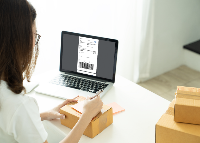 Nainen kirjoittaa muistiinpanoa paketin päälle ja käyttää Shipit-lähetystyökalua tietokoneellaan.