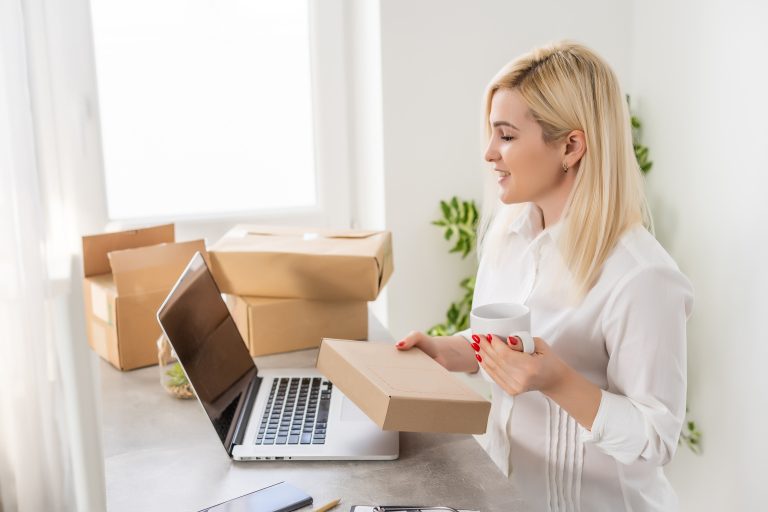 Nainen pitää kahvikuppia ja pakettia kädessään katsellen tietokoneen näyttöä kotitoimistossa.