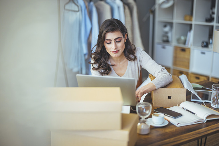 Nainen työskentelee kannettavalla tietokoneella kotitoimistossa pakettien keskellä ja valmistelee verkkokauppalähetystä.