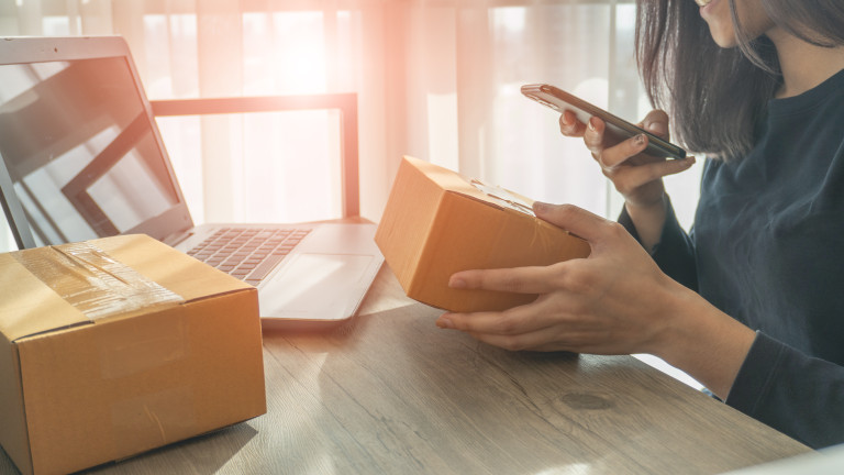Nainen tarkistaa pakettia ja käyttää älypuhelinta kannettavan tietokoneen äärellä – verkkokauppatilausten käsittelyä.