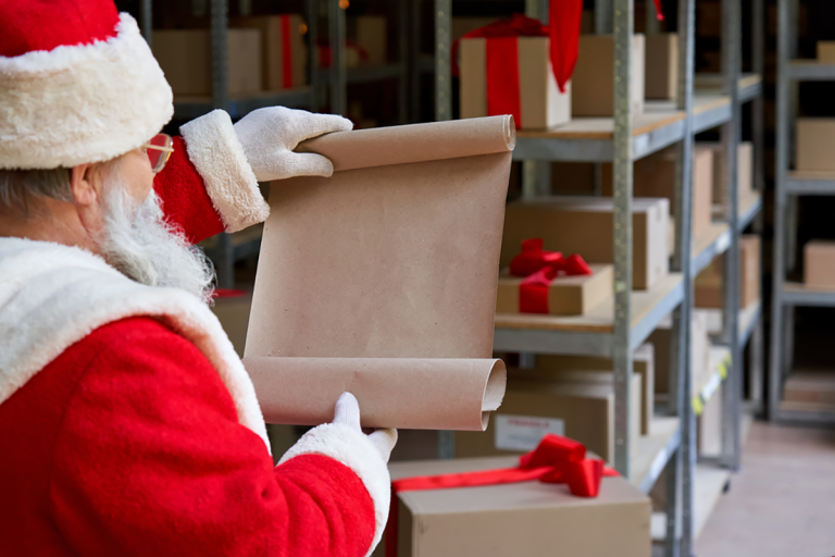 Jultomten läser en önskelista i ett lager fyllt med paket med röda band.