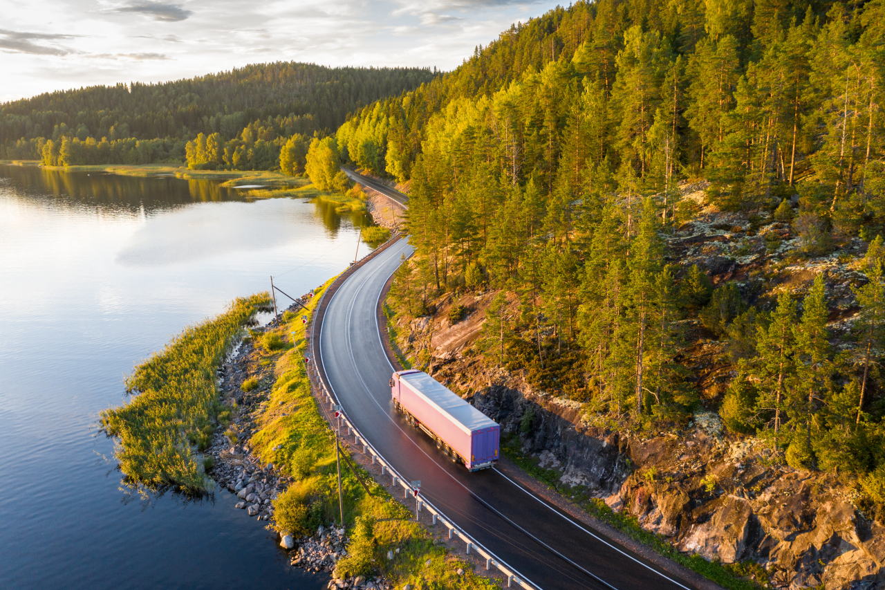 En lastbil kör längs en slingrande väg genom ett skogslandskap vid en sjö.